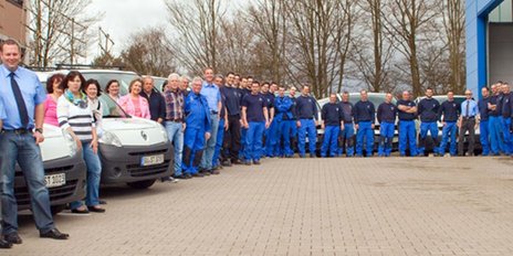 Mitarbeiter in blauen Arbeitsanzügen posieren lächelnd vor ihren Fahrzeugen, umgeben von kahlen Bäumen und einem grauen Himmel.