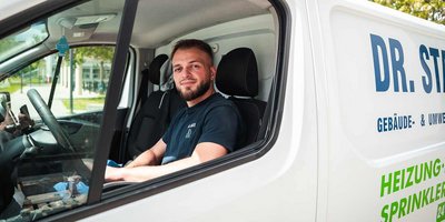 Junger Handwerker entspannt im Lieferwagen unterwegs Ein junger Mann mit Bart sitzt lächelnd im Fahrerhaus eines weißen Lieferwagens, während er sein Handy in der Hand hält.