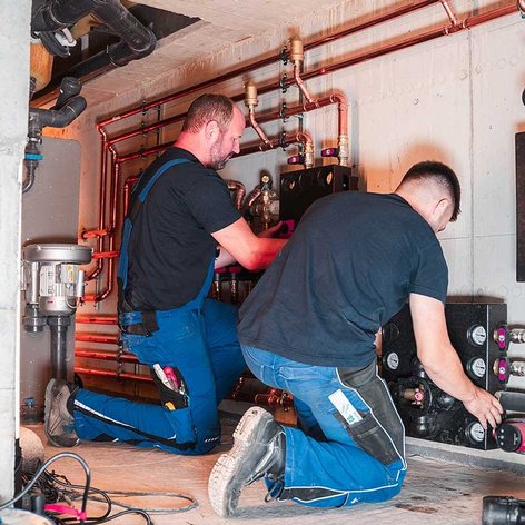 Zwei Handwerker arbeiten konzentriert an einer komplexen Rohrleitungsinstallation in einem Kellerraum.