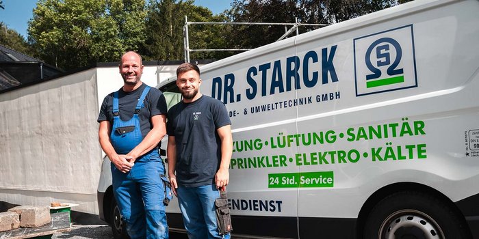 Zwei Handwerker stehen lächelnd vor einem Firmenwagen, umgeben von Baustellenmaterial und Bäumen im Hintergrund.
