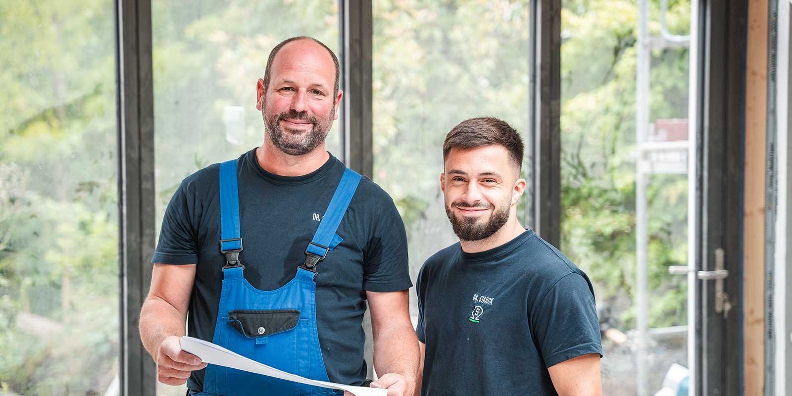 Zwei Handwerker stehen in einem hellen Raum mit großen Fenstern und lächeln, während sie Pläne in der Hand halten.